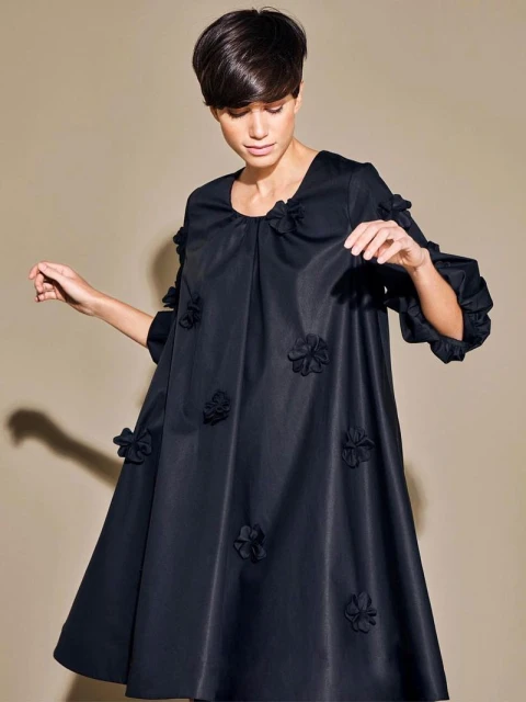 Fashionable woman in a short dark dress adorned with floral appliques posing against a neutral backdrop in a studio setting.