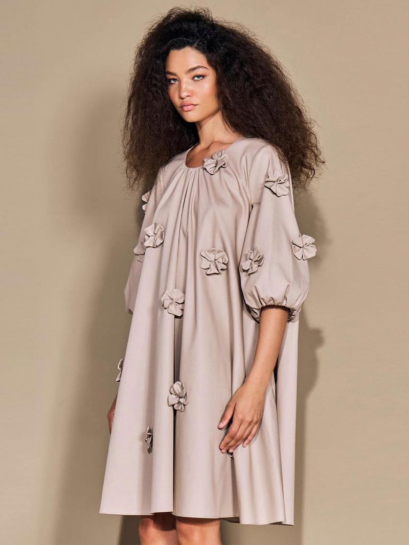 Portrait of a female fashion model in a beige dress with flower embellishments, posing in a photo studio against a neutral backdrop.