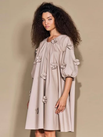 Portrait of a female fashion model in a beige dress with flower embellishments, posing in a photo studio against a neutral backdrop.
