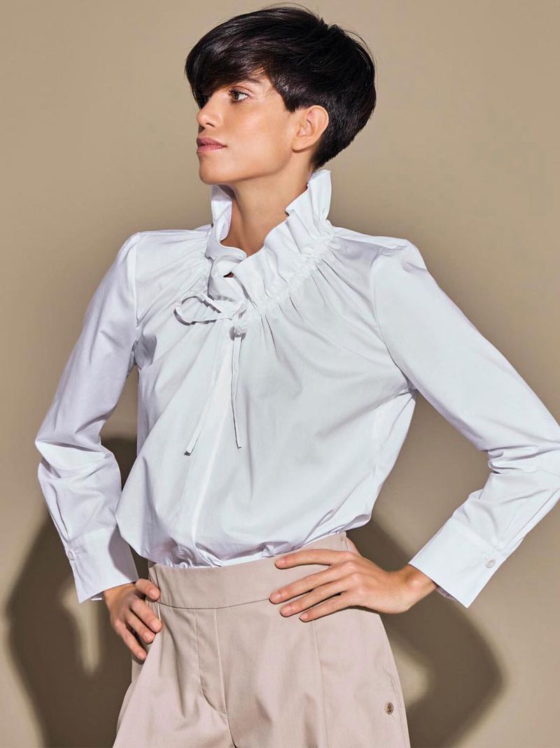 Portrait of an adult woman wearing a white ruffled blouse and khaki pants against a neutral background in a studio shot.