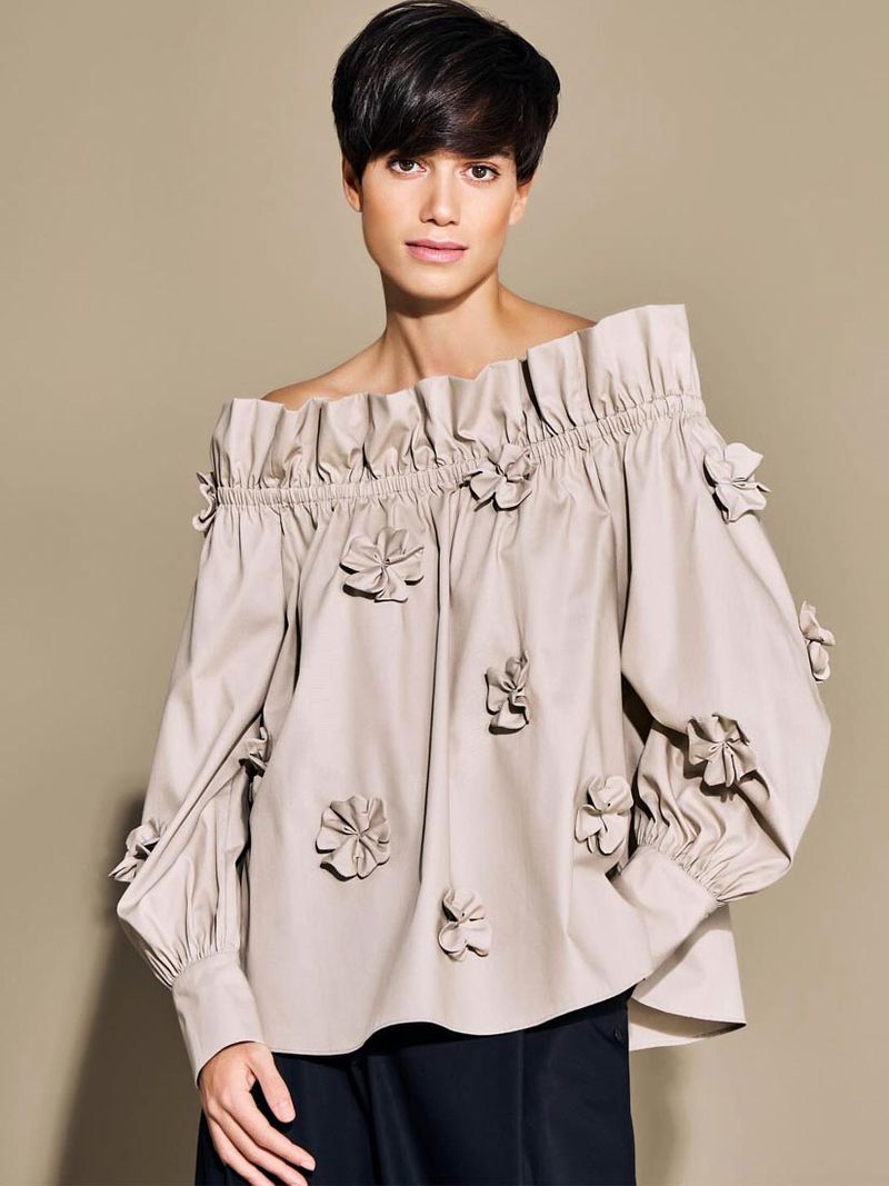 A woman in an off-the-shoulder blouse poses in front of a neutral background, showcasing fashion and style with a modern edge.