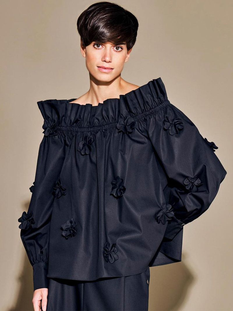 An adult woman with short dark hair poses in a studio wearing a black floral off-the-shoulder blouse, in a fashion studio setting.