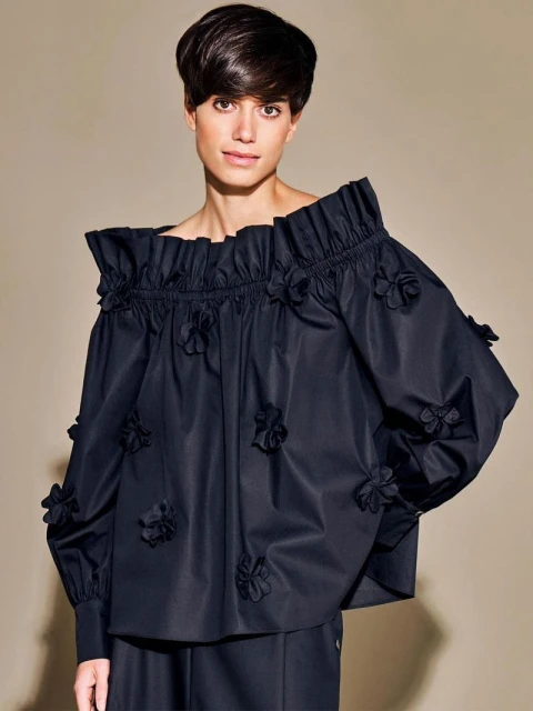 An adult woman with short dark hair poses in a studio wearing a black floral off-the-shoulder blouse, in a fashion studio setting.