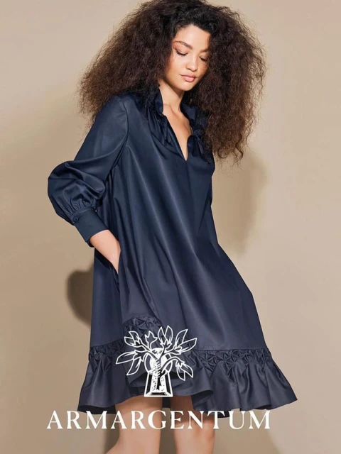 A woman with curly hair models a navy blue dress with long sleeves in a studio setting, showcasing fashion and style.