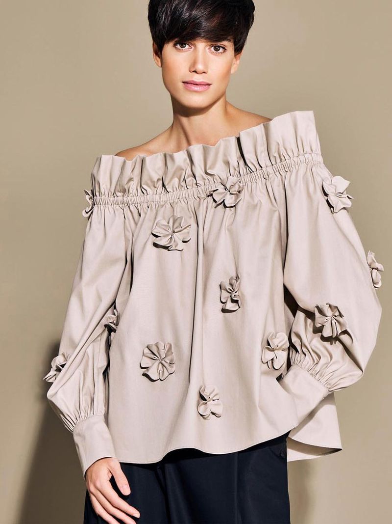 Fashion portrait of a woman wearing an off-the-shoulder blouse with flower details, looking at the camera with a neutral expression.