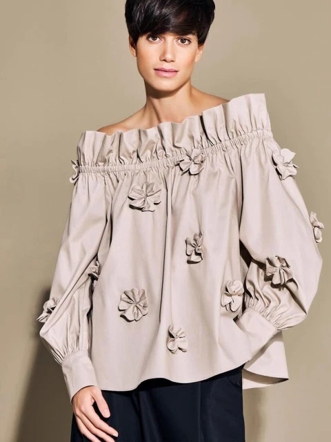 Fashion portrait of a woman wearing an off-the-shoulder blouse with flower details, looking at the camera with a neutral expression.