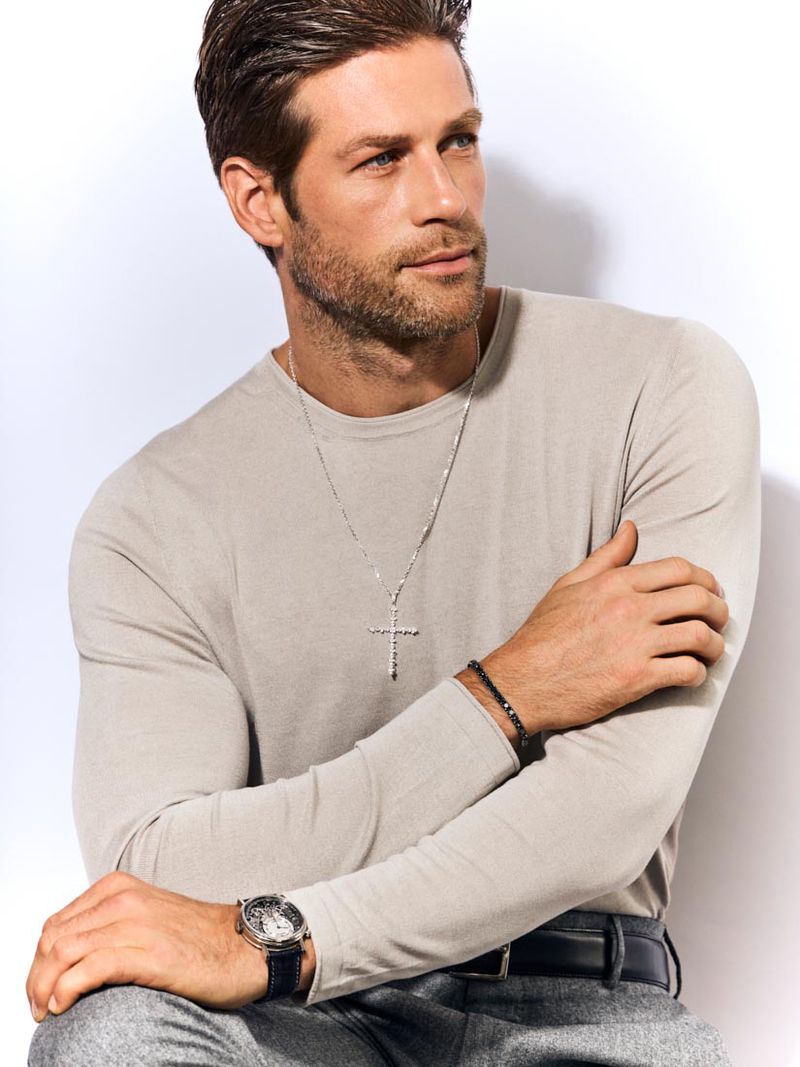 Adult male poses indoors wearing a beige long-sleeve shirt with grey pants, wristwatch, bracelets, and necklace with a cross pendant.