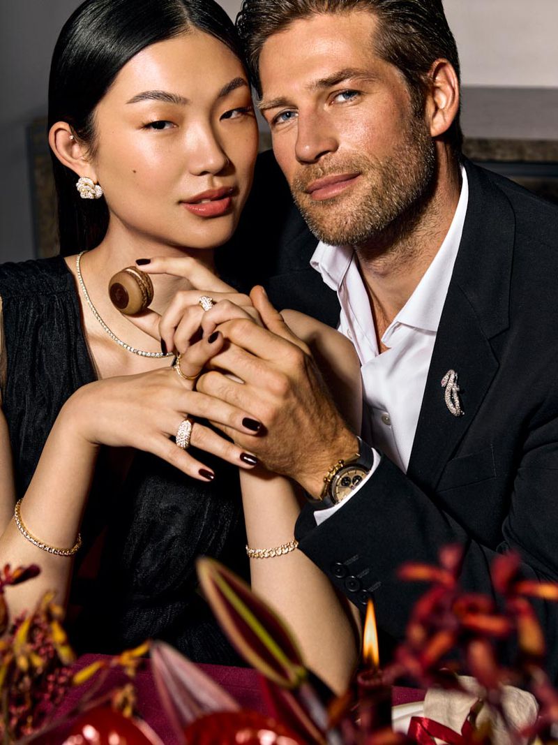 Elegant portrait of an Asian woman and Caucasian man in formal attire, posing intimately with jewelry, against a dark, romantic backdrop.