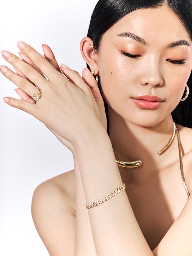 Close-up of a young Asian woman wearing gold jewelry, including a ring, earrings, bracelet, and necklace, posing against a white backdrop.