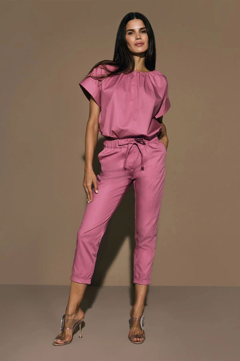 A full shot of a brunette woman in a pink pantsuit and silver high heels posing against a neutral brown background in a studio setting.