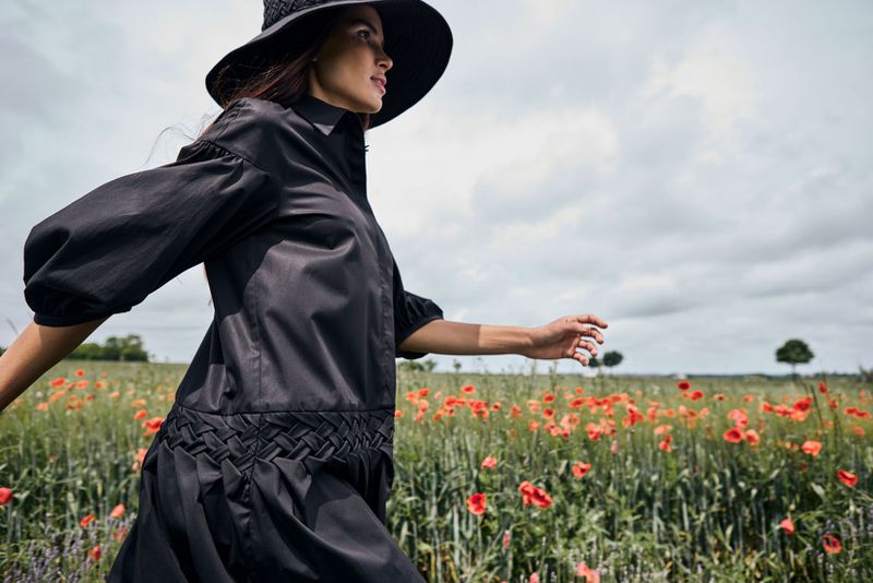 Model Raica Oliveira walking through a field of red poppies wearing a black dress and wide-brim hat