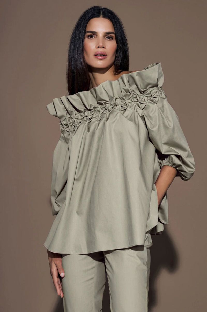 A brunette woman poses in a studio with a neutral background, wearing a tan, off-the-shoulder blouse and matching pants.