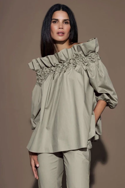 A brunette woman poses in a studio with a neutral background, wearing a tan, off-the-shoulder blouse and matching pants.