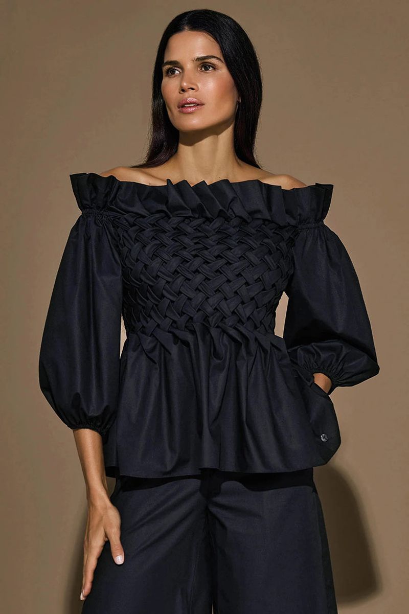 A brunette woman poses in a black, off-the-shoulder woven top and matching pants against a neutral backdrop in a fashion shot.