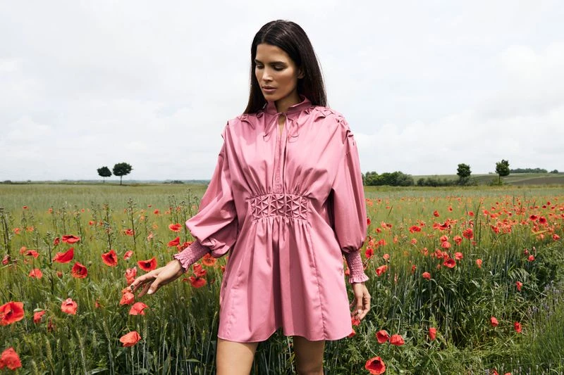 A woman in a pink dress standing in a field of red poppies on a cloudy day. The dress has long sleeves and a gathered waist.
