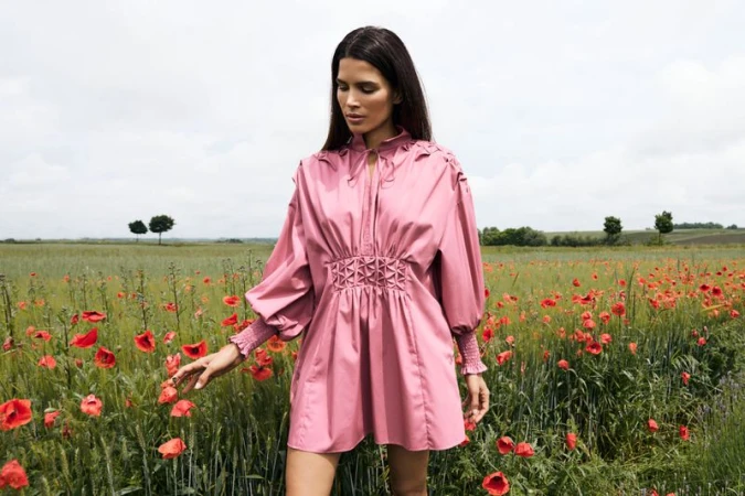 A woman in a pink dress standing in a field of red poppies on a cloudy day. The dress has long sleeves and a gathered waist.