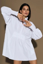 Model wearing a loose white dress with long puffed sleeves posing against a plain brown background