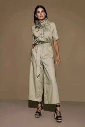 Model Raica Oliveira wearing a beige jumpsuit with a ruffled collar and platform wedge sandals against a warm neutral studio background.