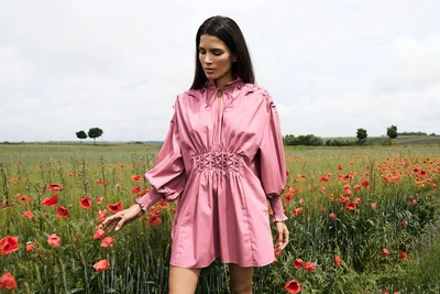 Model wearing a pink smocked dress standing in a green meadow filled with red poppy flowers under an open sky