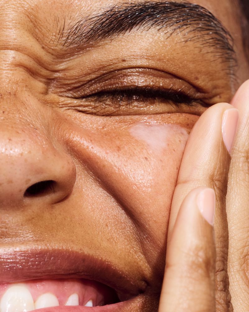 Close-up of an adult female with dark skin smiling and touching her face, highlighting skincare and beauty with natural lighting.