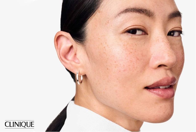 Close-up studio shot of an adult Asian woman with freckles, hoop earrings, and a white turtleneck sweater against a white backdrop.