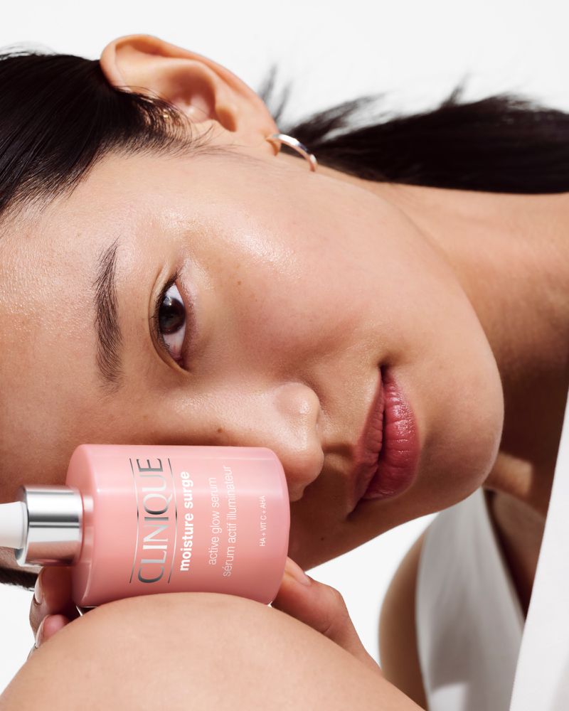 Close-up shot of an Asian woman with fair skin holding a pink Clinique Moisture Surge bottle against a bright, neutral background.