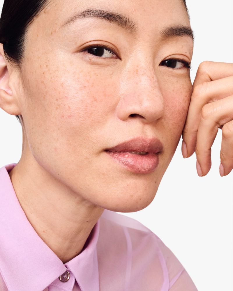 Close-up portrait of a young Asian woman with light skin and freckles, wearing a light pink button-down shirt against a white background.