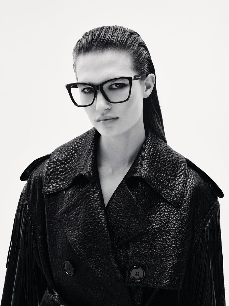 Close-up studio shot of a woman wearing a dark trench coat and square-frame eyeglasses against a plain white background.