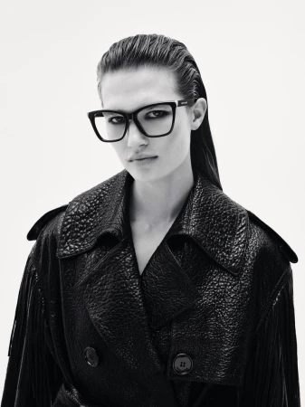 Close-up studio shot of a woman wearing a dark trench coat and square-frame eyeglasses against a plain white background.