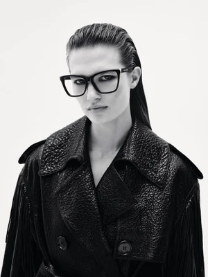 Model wearing black eyeglasses and a textured black trench coat against a solid white background
