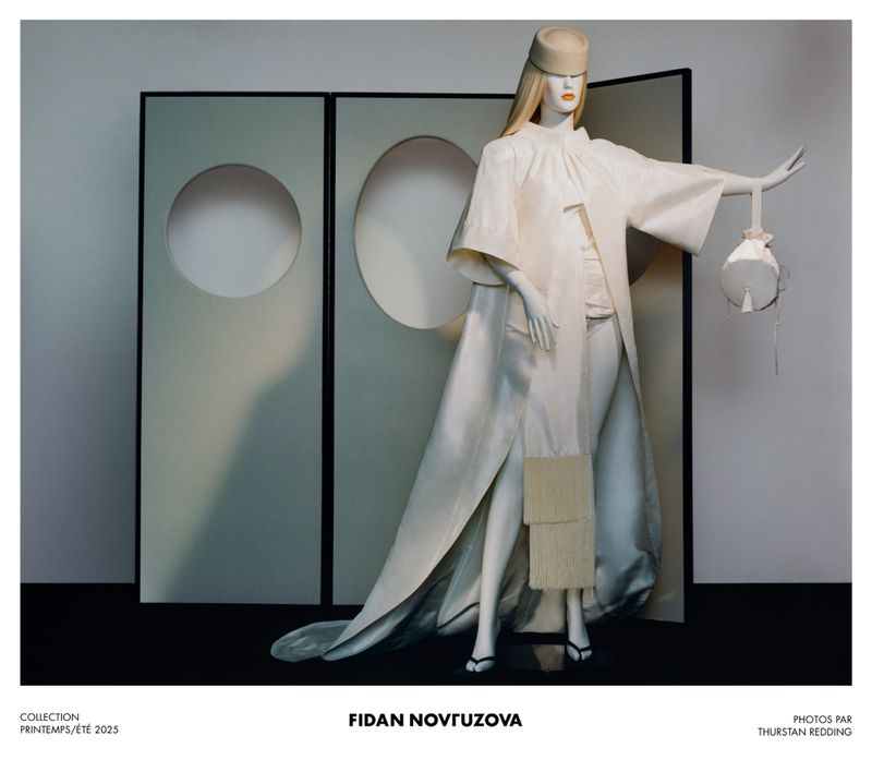 Mannequin displays an elegant off-white ensemble featuring a long coat and handbag, against a backdrop of panels with circular cutouts.