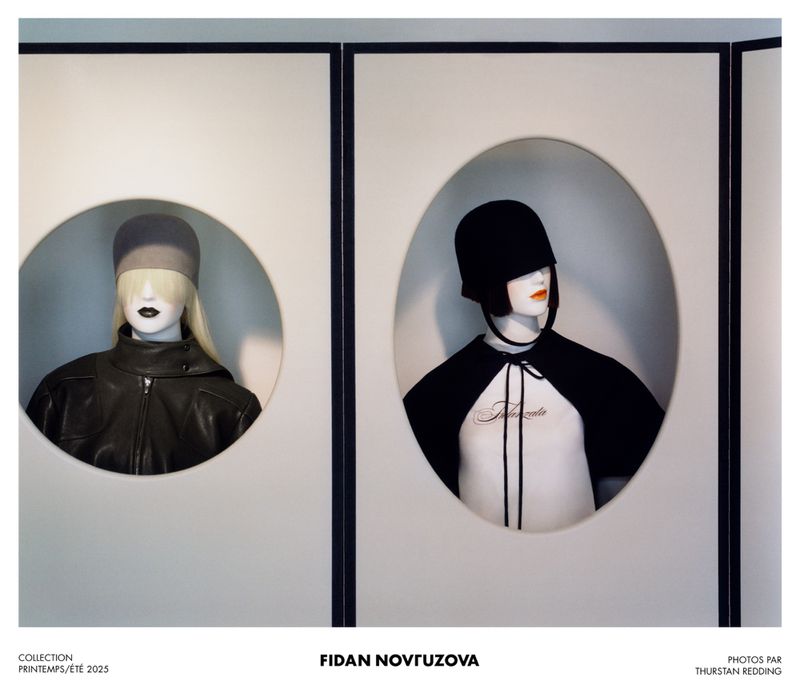 Two mannequins wearing black and gray hats and jackets, and a black cape with a white shirt, displayed in a minimalist showroom.