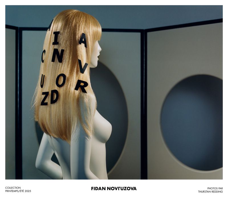 Mannequin headshot with a blonde wig adorned with black letters, in front of a light blue and beige geometric patterned screen.