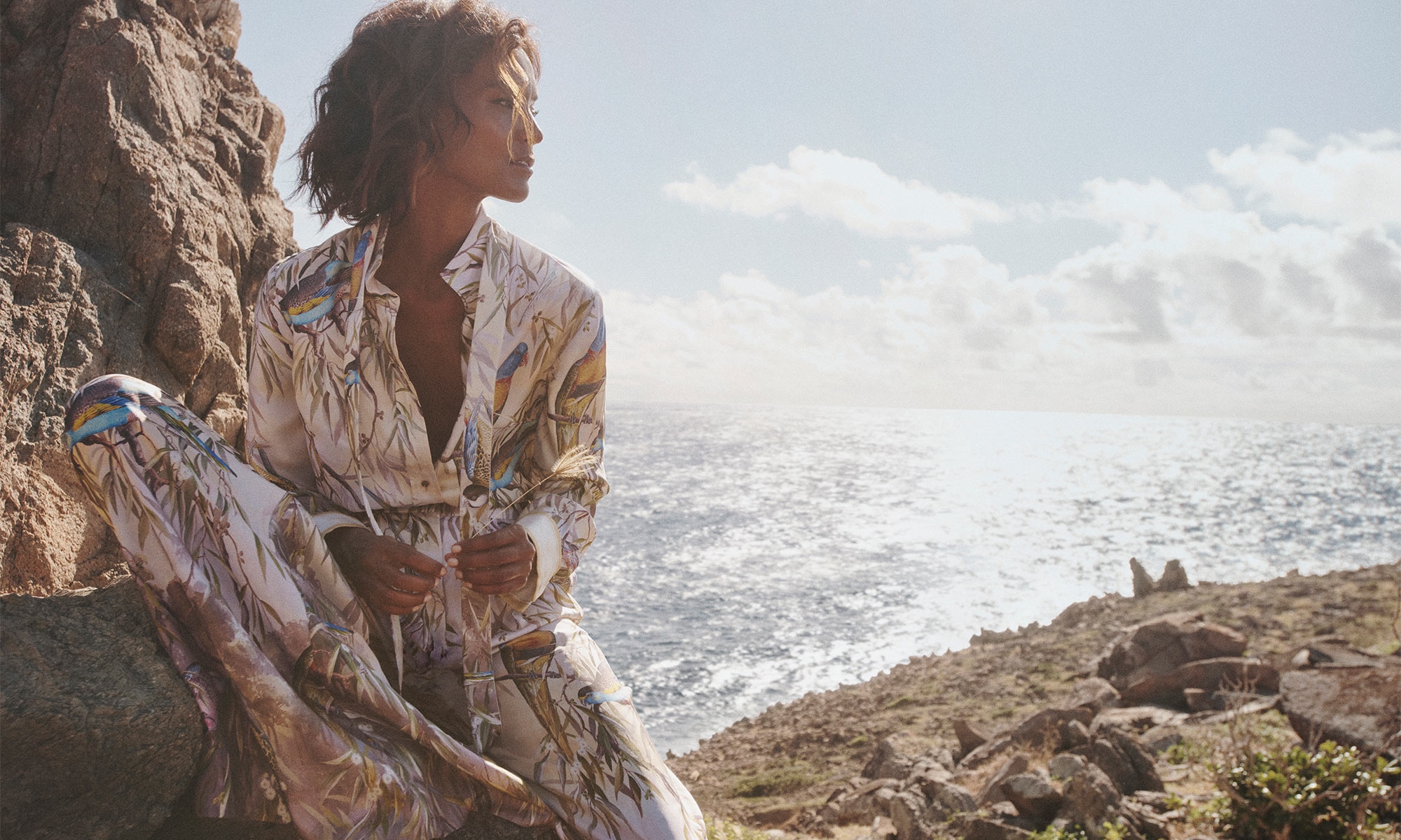 Brunette adult woman in a patterned pajama-style outfit sitting on rocks near the ocean on a partly cloudy day, looking toward the horizon.