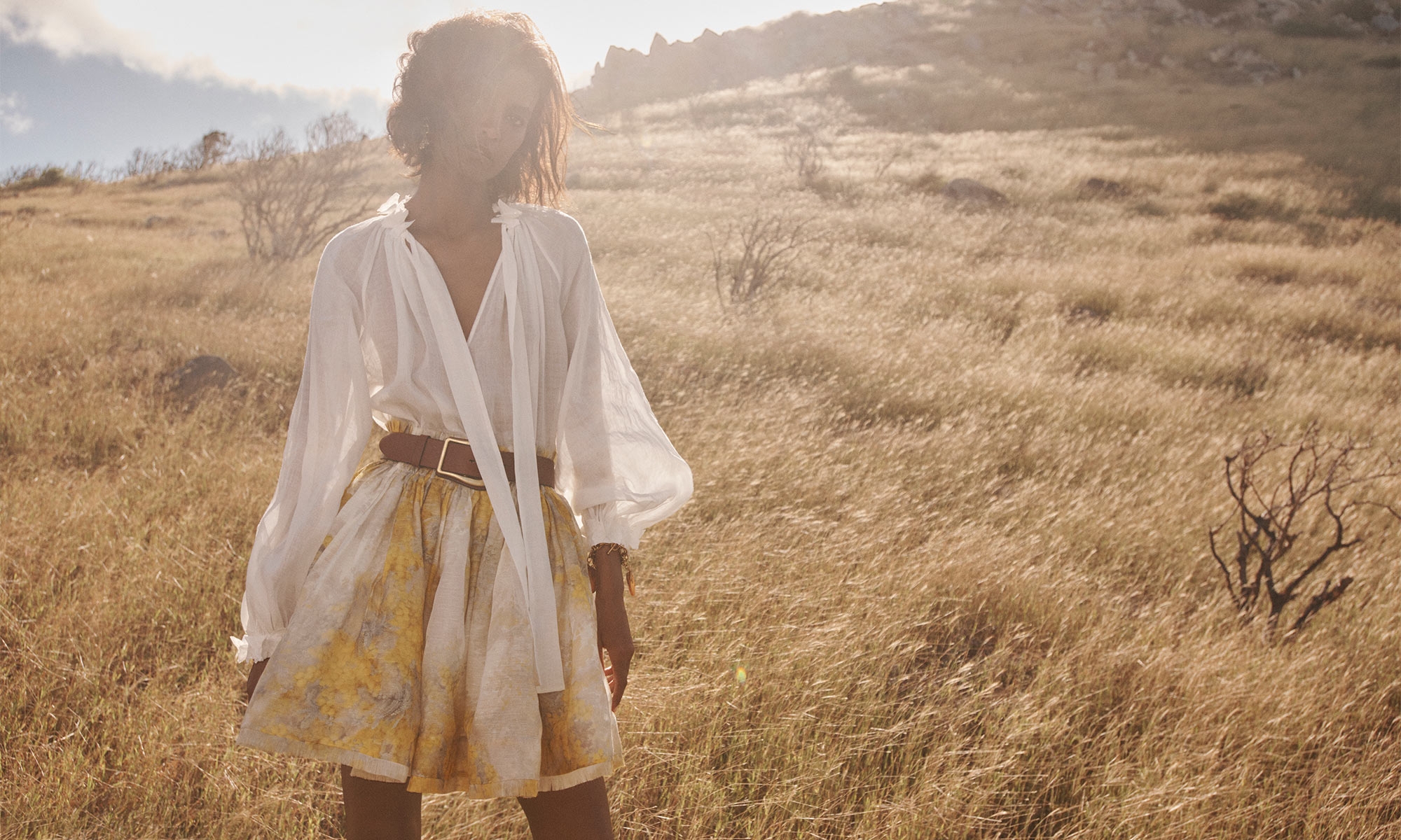 Woman wearing a white blouse and yellow skirt poses outdoors in a sunlit field of golden grass. Fashion, style, and nature.