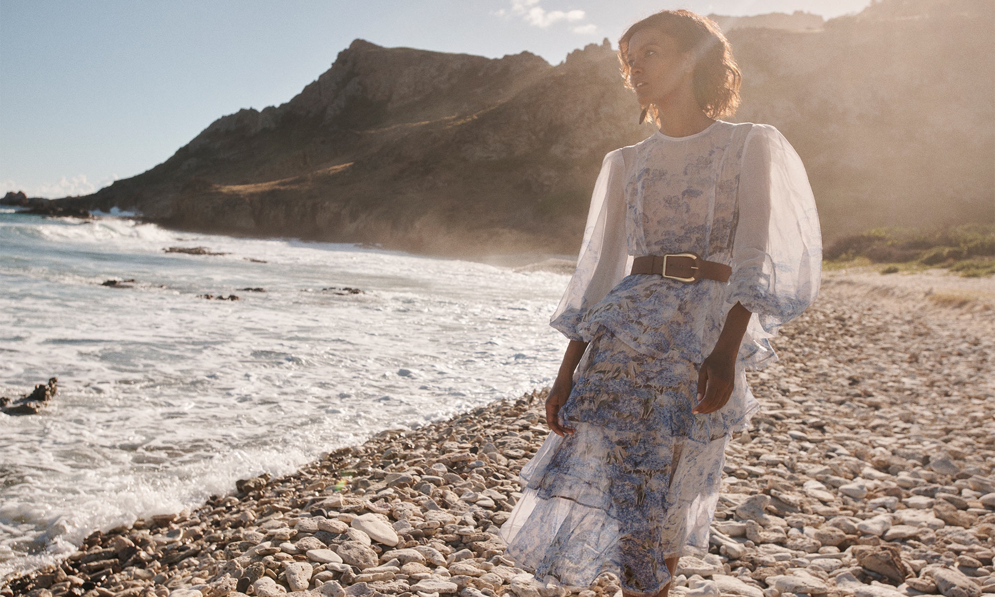A woman poses on a rocky beach in a blue floral dress, with a belt and a mountain and ocean in the background, bathed in sunlight.