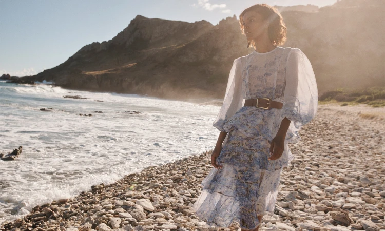A woman poses on a rocky beach in a blue floral dress, with a belt and a mountain and ocean in the background, bathed in sunlight.