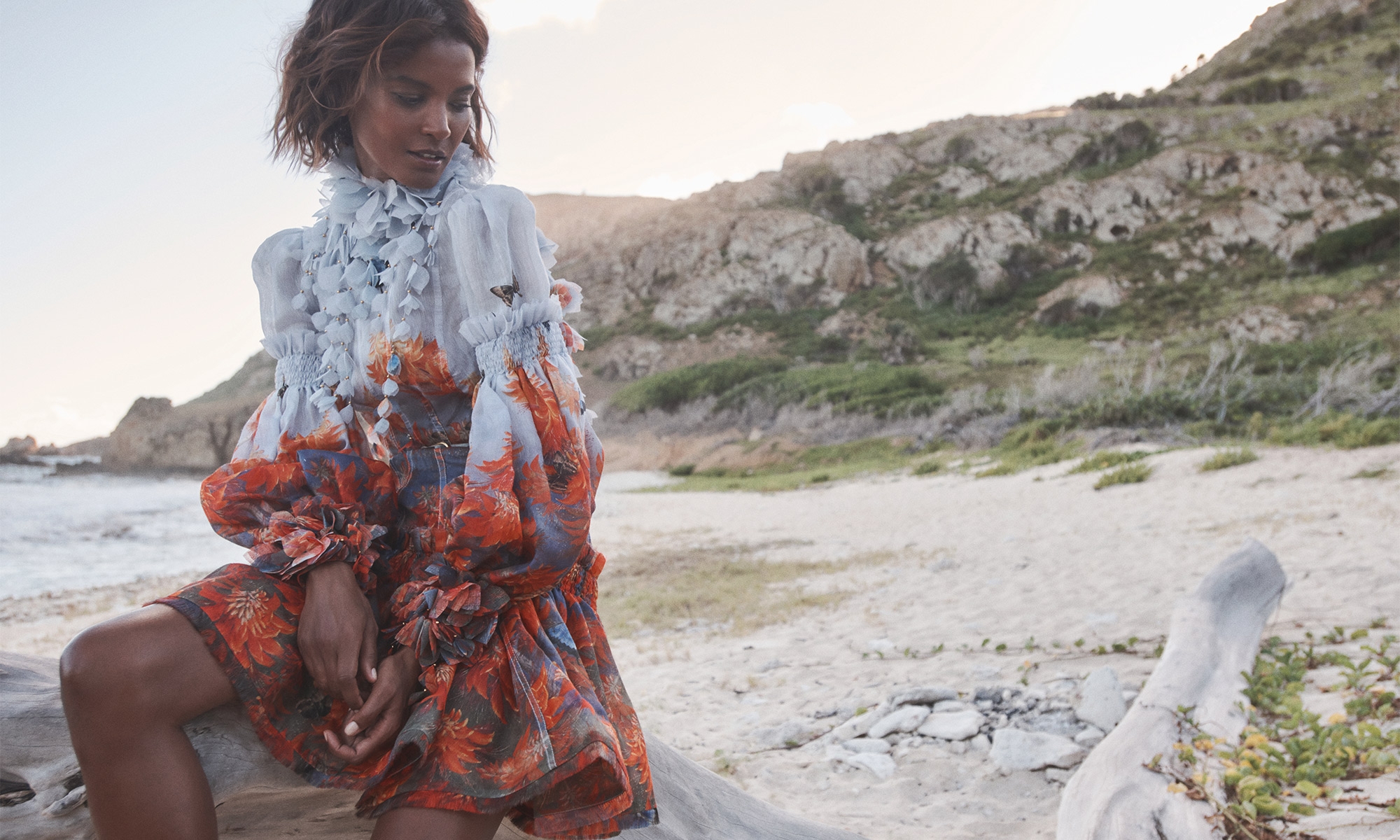 Woman in floral dress poses on driftwood at beach with blue sky and hilly horizon; fashion, nature and coastal themes, outdoor setting.