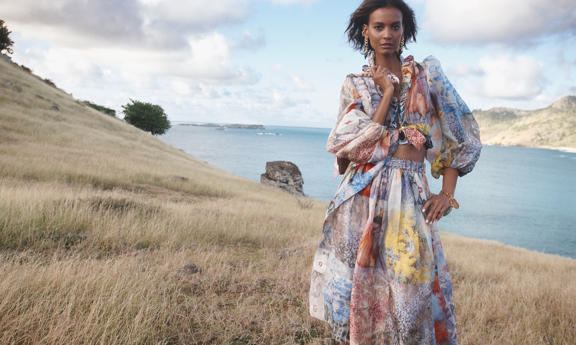 A woman in a patterned dress stands on a grassy hill overlooking the ocean under a partly cloudy sky in a fashion editorial.