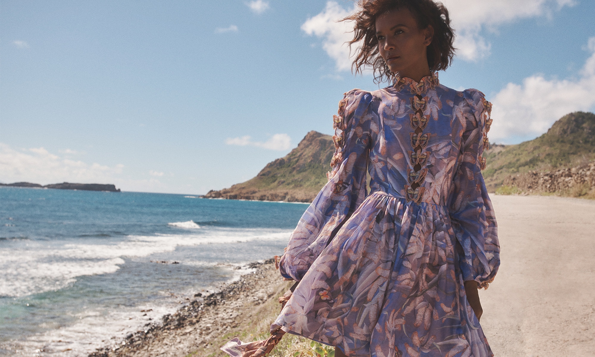 A tanned-skinned model poses in a flowing floral dress against a blue ocean and sky backdrop, epitomizing elegance and natural beauty.