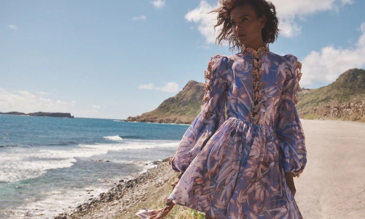 A tanned-skinned model poses in a flowing floral dress against a blue ocean and sky backdrop, epitomizing elegance and natural beauty.