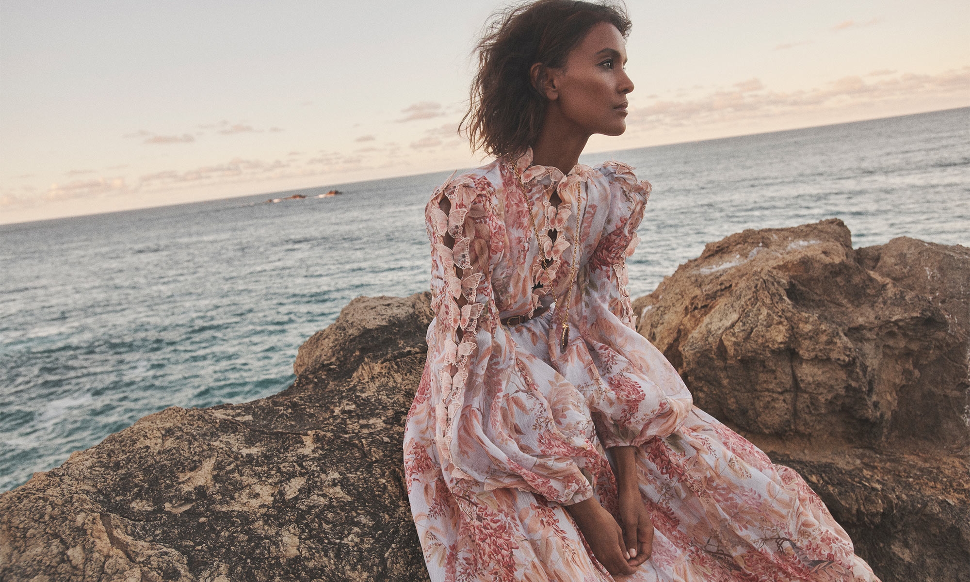 Model Liya Kebede seated on rocky coast wearing printed ruffle dress for Zimmermann Spring RTW campaign.