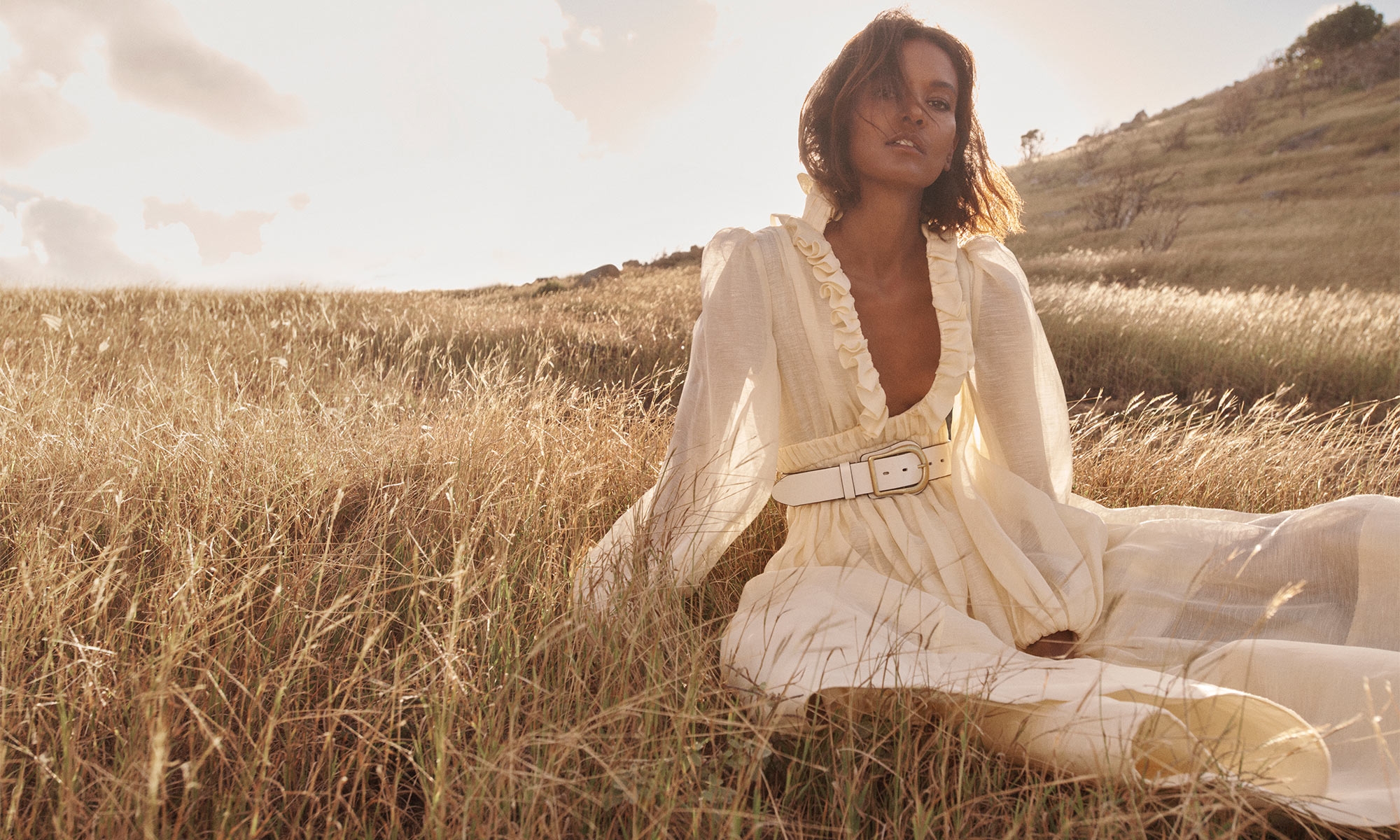 Liya Kebede seated in tall dry grass wearing a cream-colored belted dress under bright sunlight for Zimmermann Spring RTW