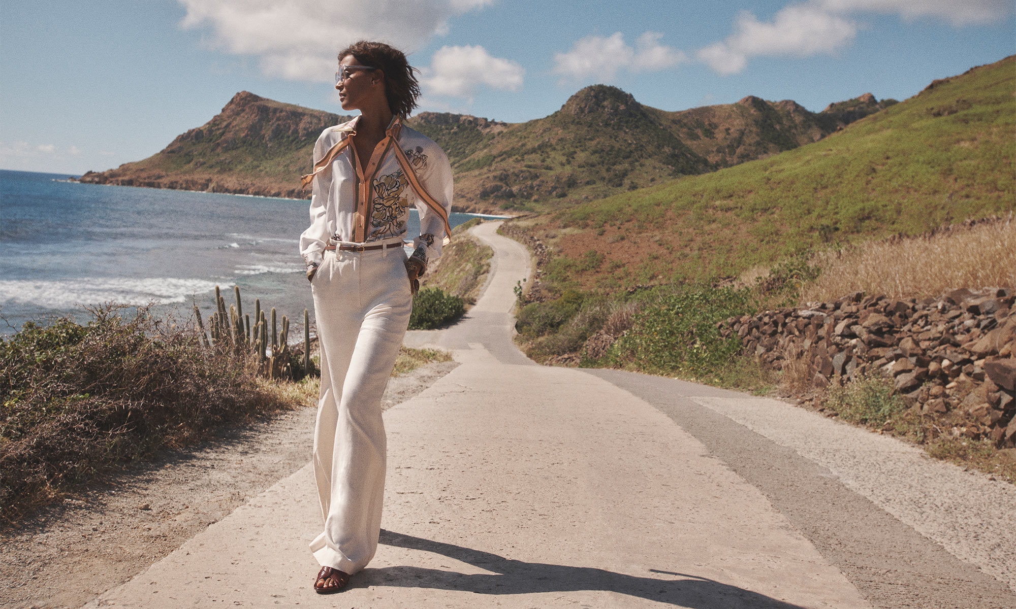 A woman walks on a road with hills and water views in the background. She wears a white patterned shirt and pants outdoors.