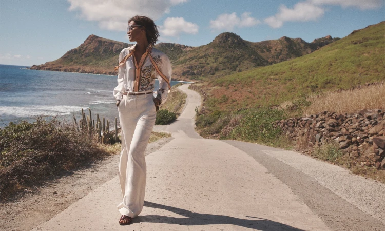 A woman walks on a road with hills and water views in the background. She wears a white patterned shirt and pants outdoors.
