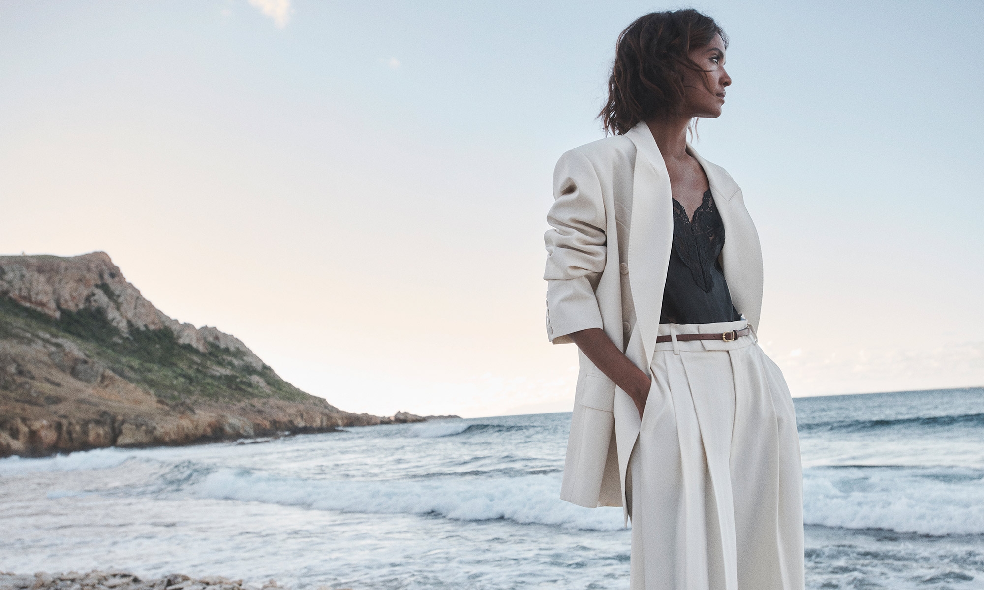 Model Liya Kebede standing on a rocky coast wearing a cream suit jacket and trousers for Zimmermann Spring RTW