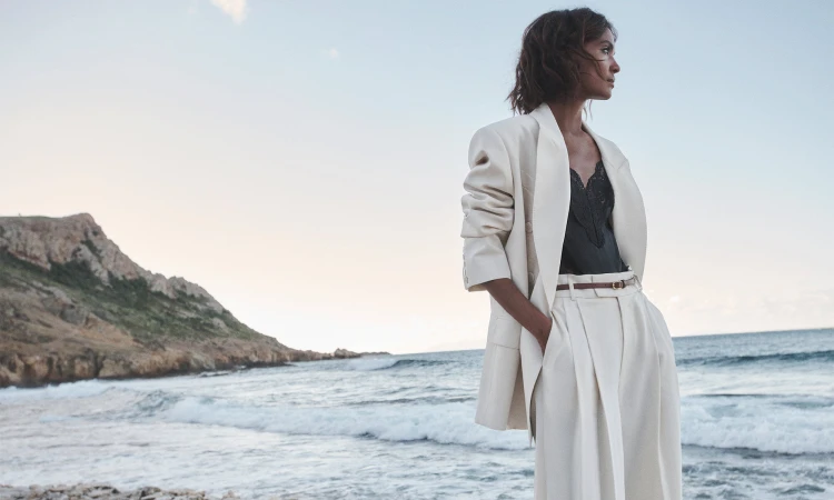 Model Liya Kebede standing on a rocky coast wearing a cream suit jacket and trousers for Zimmermann Spring RTW
