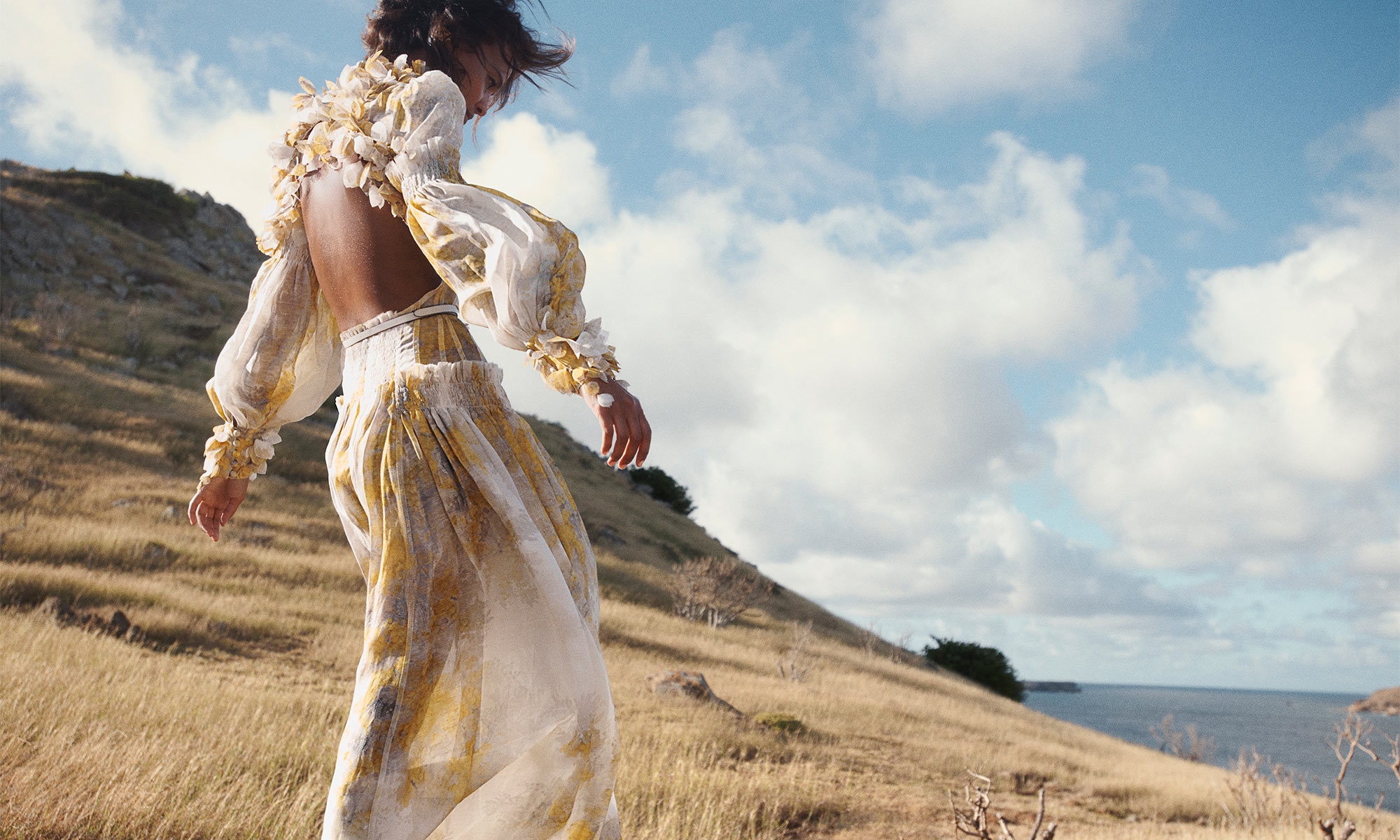 An adult woman stands in an open back yellow dress on a grassy hillside on a sunny day with a blue sky filled with white clouds.