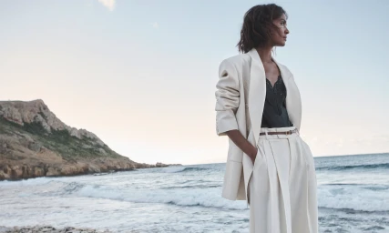 Model Liya Kebede standing on a rocky coast wearing a cream suit jacket and trousers for Zimmermann Spring RTW