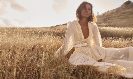 Liya Kebede seated in tall dry grass wearing a cream-colored belted dress under bright sunlight for Zimmermann Spring RTW